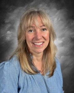 Smiling woman with long blonde hair wearing a light blue blouse, set against a soft gray background.
