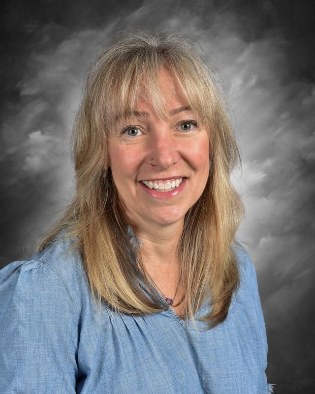 Smiling woman with long blonde hair wearing a light blue blouse, set against a soft gray background.