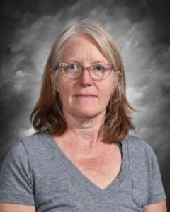 Portrait of a mature woman with shoulder-length hair, wearing a gray t-shirt, and glasses, against a soft gray background. The image captures her calm demeanor and thoughtful expression.