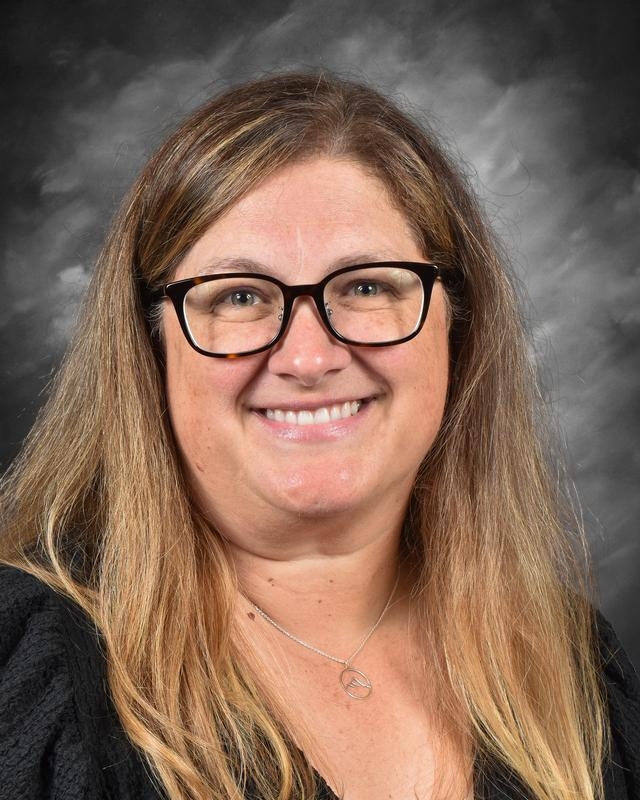 Professional headshot of a woman with long, straight brown hair, wearing black glasses and a black top, smiling against a dark, textured background.