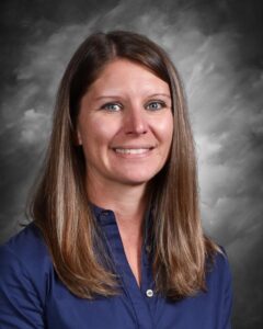 Portrait of a smiling woman with long brown hair, wearing a blue shirt, set against a gray background. The image conveys a professional and approachable demeanor, suitable for a staff profile or educational context.