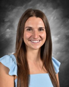 Smiling young woman with long brown hair wearing a light blue dress, set against a dramatic gray background.