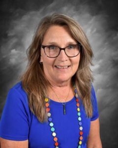 Portrait of a woman with long, wavy hair and glasses, wearing a blue shirt and a colorful beaded necklace, set against a gray background.