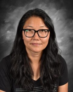 Portrait of a woman with long black hair and glasses, wearing a black T-shirt with a striped design, set against a gray background.