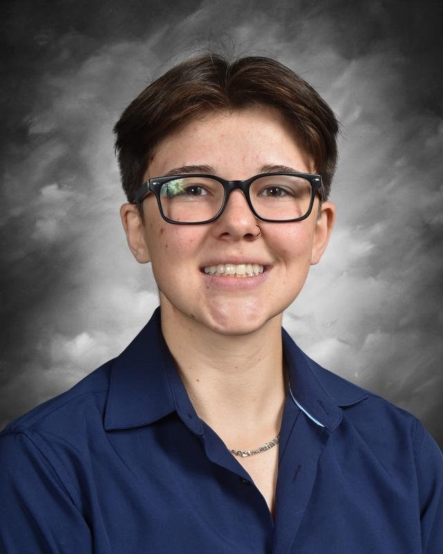 Portrait of a young individual with short hair and glasses, smiling confidently while wearing a blue collared shirt against a soft gray background.