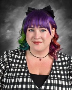 Portrait of a woman with vibrant rainbow-colored hair, styled in loose curls. She wears a black and white checkered top and a black bow in her hair, accessorized with earrings and a choker necklace. The background is a soft gray, highlighting her cheerful expression and unique style.