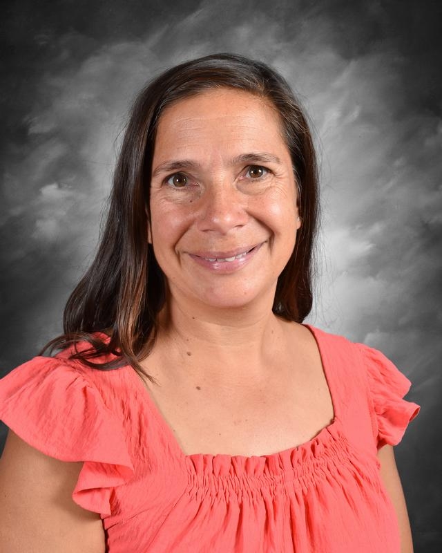 Smiling woman with long dark hair wearing a coral-colored top, set against a gray background. Ideal for showcasing friendly and approachable personality in professional or educational contexts.
