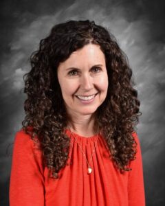 Smiling woman with curly hair wearing an orange top, posing against a gray backdrop.