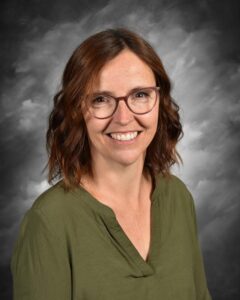 Smiling woman with shoulder-length brown hair and glasses, wearing a green blouse, against a gray background. Ideal for professional profiles or educational contexts.