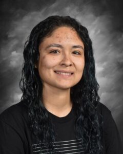Portrait of a young woman with long, wavy black hair, wearing a black t-shirt with a striped design, smiling against a gray backdrop.