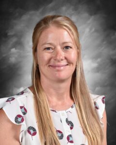 Portrait of a smiling woman with long blonde hair, wearing a patterned white blouse, against a grey background. Ideal for profiles or team introductions.