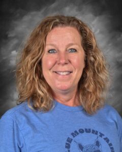 Professional headshot of a smiling woman with curly hair, wearing a blue t-shirt featuring the text "Blue Mountain." The background is a soft gray gradient, enhancing her friendly expression.