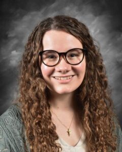 Young woman with curly hair and glasses smiling in a portrait against a gray background, wearing a light sweater and a necklace.