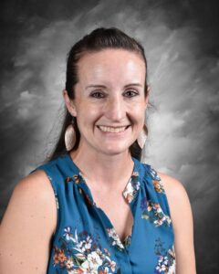 Smiling woman with long brown hair wearing a blue floral blouse and large earrings, posing in front of a gray backdrop. Ideal for a professional profile or team introduction.