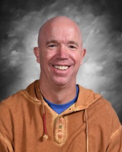 Smiling man with a bald head wearing a brown hoodie over a blue shirt, set against a soft gray background.