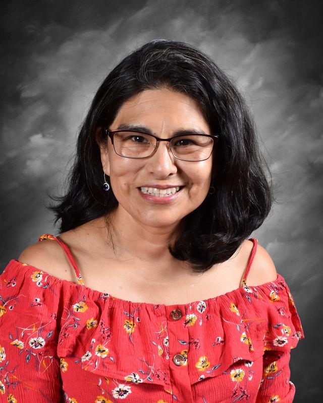 Portrait of a smiling woman with long black hair, wearing glasses and a red floral off-the-shoulder top, set against a soft gray background.