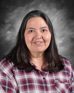 Portrait of a woman with long dark hair, wearing a red and white plaid shirt, smiling against a gray background.