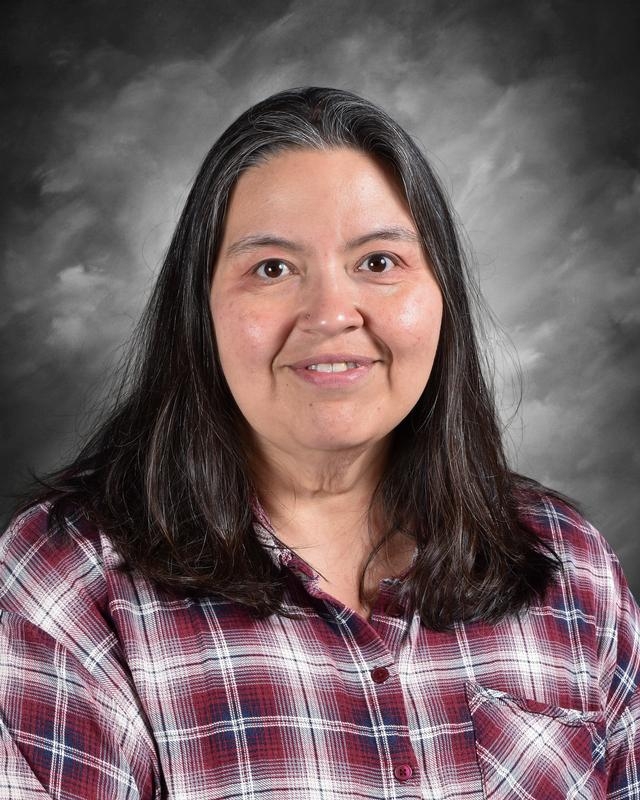 Portrait of a woman with long dark hair, wearing a red and white plaid shirt, smiling against a gray background.