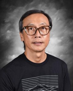 Portrait of a middle-aged man wearing glasses and a black t-shirt with a mountain graphic, set against a textured gray background. The man has short black hair and a slight smile, conveying a friendly demeanor.