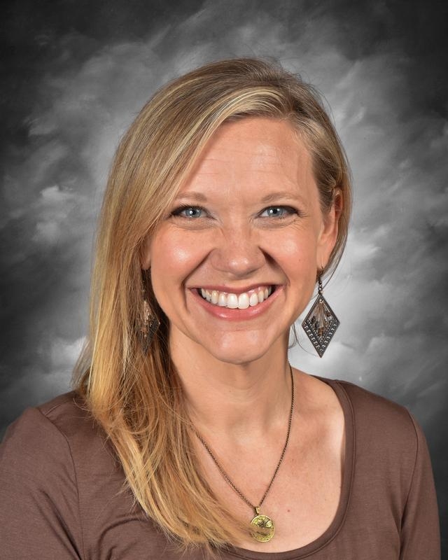 Smiling woman with long blonde hair wearing a brown shirt and decorative earrings, posing against a gray background.