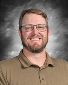 Smiling man with glasses and a beard, wearing a brown polo shirt, against a dramatic gray background. Ideal for profiles or professional contexts.