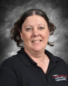 Portrait of a woman with shoulder-length hair styled in curls, wearing a black polo shirt with the logo of St. Rain Valley Schools, smiling against a gray backdrop.