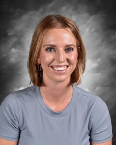 Smiling young woman with short blonde hair, wearing a gray shirt, against a blurred gray background.
