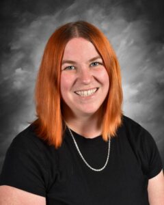 Smiling woman with shoulder-length orange hair wearing a black shirt and silver necklace, posing against a gray backdrop.
