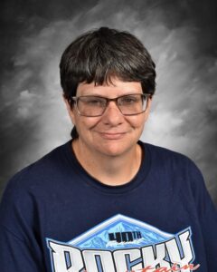Portrait of a woman with short, dark hair and glasses, wearing a blue "Rocky Mountain" graphic t-shirt, set against a gray backdrop.