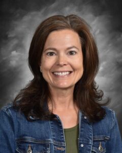 Smiling woman with long brown hair wearing a denim jacket over a green top, set against a gray background. Ideal for showcasing approachable and friendly personas in professional settings.