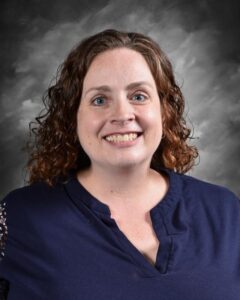 Portrait of a woman with curly hair wearing a navy blue top, smiling against a gray backdrop. Ideal for profiles or team introductions.