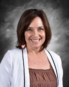 Portrait of a smiling woman with shoulder-length brown hair, wearing heart-shaped earrings and a white cardigan over a brown top, set against a gray backdrop.