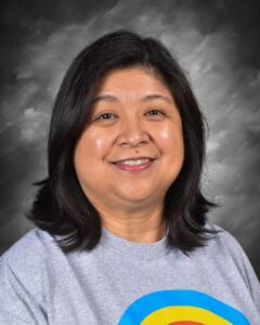 Smiling woman with long black hair wearing a gray t-shirt featuring a colorful design, set against a dark gray background.