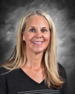 Smiling woman with long blonde hair, wearing a black v-neck shirt, posing against a blurred gray background. Ideal for showcasing friendly and approachable personality in professional settings.