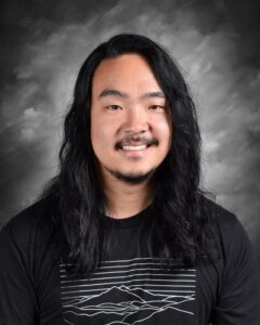 Portrait of a smiling man with long black hair, wearing a black t-shirt featuring a mountain graphic, against a soft gray background.