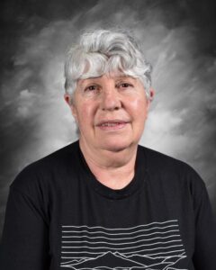 Portrait of an older woman with gray hair, wearing a black t-shirt with a graphic design, set against a gray background. She has a gentle expression, conveying warmth and approachability.