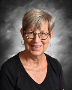 Smiling older woman with short gray hair and glasses, wearing a black shirt, against a gray background.