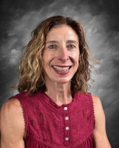 Smiling woman with curly hair wearing a burgundy sleeveless top, posing against a gray backdrop. The image captures her friendly expression, highlighting her enthusiasm and approachability.