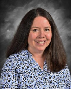 Portrait of a woman with long dark hair, smiling, wearing a blue and black patterned blouse, set against a gray background. Ideal for professional profiles or educational contexts.