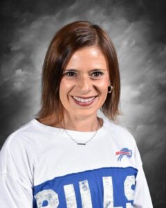 Smiling woman with short brown hair wearing a white t-shirt featuring the Buffalo Bills logo, against a gray background.