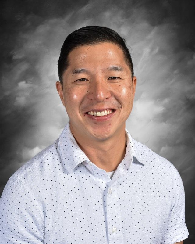 Smiling man with short dark hair wearing a white button-up shirt with blue polka dots, set against a gray background.