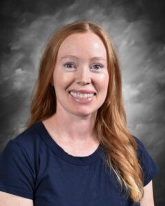 Portrait of a smiling woman with long red hair, wearing a navy blue shirt, against a gray background. Ideal for showcasing personal profiles or professional introductions.