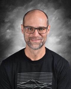 Portrait of a smiling man with glasses and a beard, wearing a black t-shirt featuring a mountain graphic, set against a soft, cloudy background.