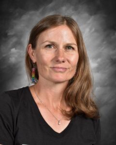 Portrait of a woman with long brown hair wearing colorful earrings and a black shirt, against a gray background. She has a neutral expression and is looking directly at the camera.
