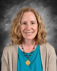 Smiling woman with curly hair wearing a teal shirt and a beige cardigan, accessorized with a colorful beaded necklace, against a gray backdrop.