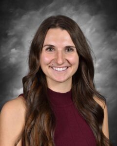 Smiling woman with long brown hair wearing a maroon sleeveless top, set against a textured gray background.