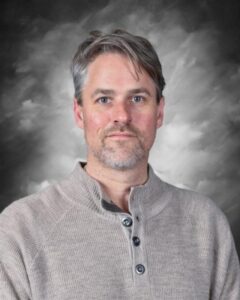 Portrait of a man with short gray hair and a beard, wearing a light gray sweater, against a dark, cloudy background. The image conveys a professional and approachable demeanor.